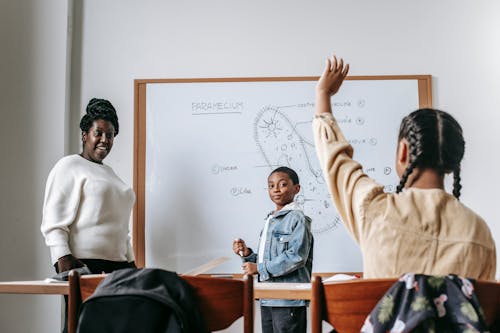 Ethnic girl raising hand while African American female teacher standing near whiteboard with teen boy and explaining task