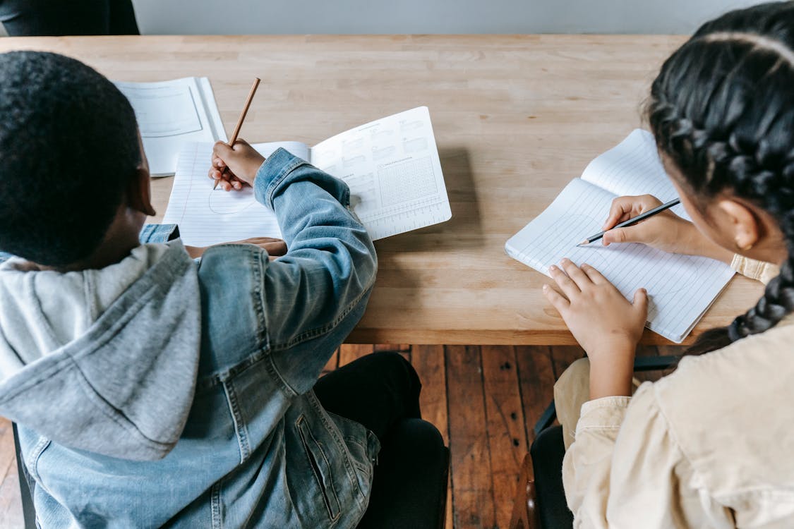 High angle of faceless ethnic schoolchildren in casual clothes sitting at wooden desk with notebooks and pencils while writing task during lesson