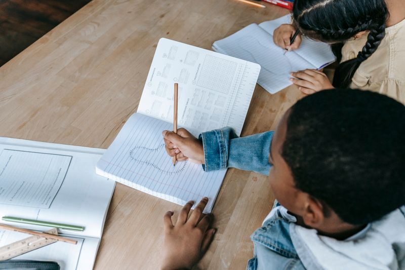 From above of crop unrecognizable students in casual clothes sitting at desk with copybook and doing exercise in classroom