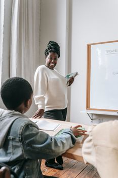 African American female teacher in casual clothes holding books while standing near whiteboard and looking at schoolboy during lesson