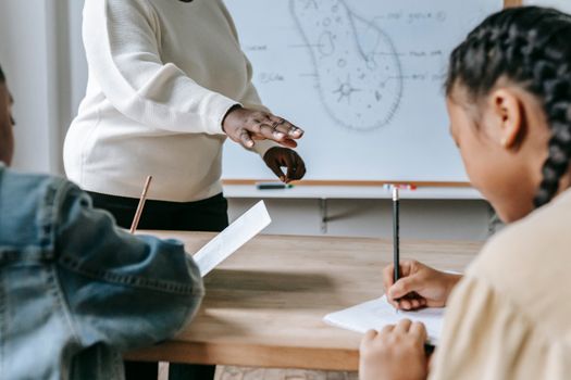 Crop faceless female teacher standing near desk while schoolchildren writing task on papers during lesson