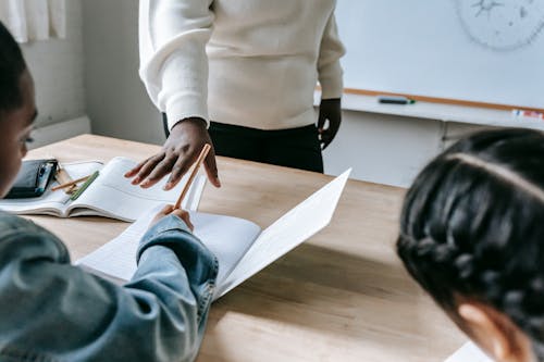 Crop faceless woman standing near desk while schoolchildren writing test with pencils and copybooks in classroom