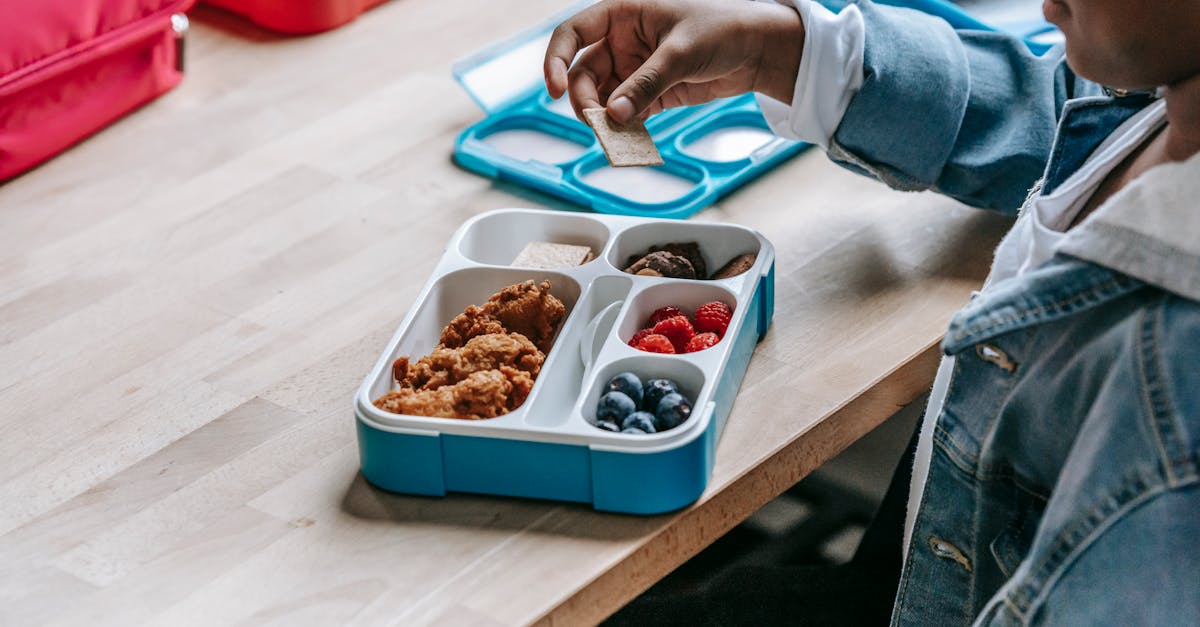 From above side view of crop unrecognizable ethnic schoolkid at table with lunch container full of tasty food