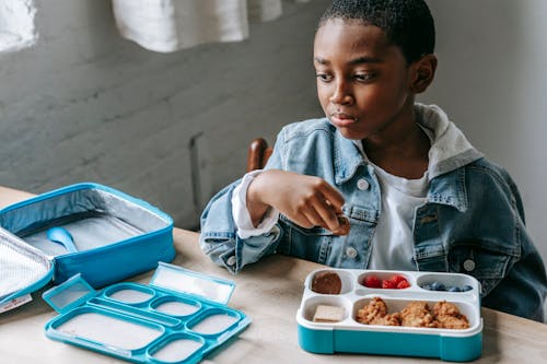 Crop contemplative African American schoolchild looking away at table with lunch container full of yummy food