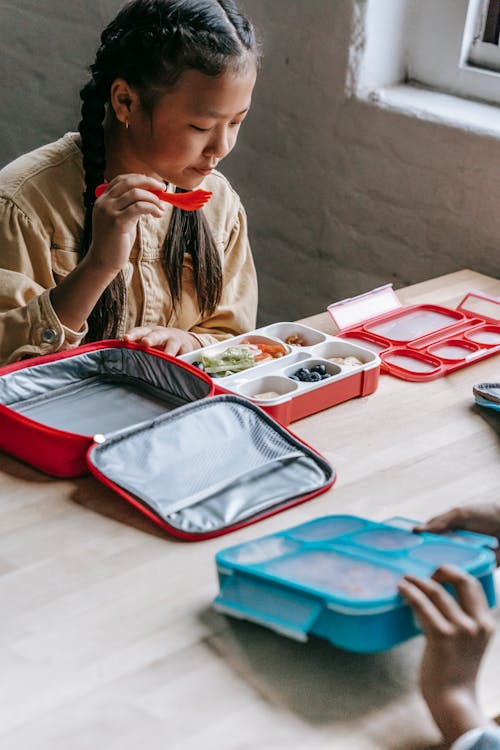 Crop adorable ethnic girl choosing healthy food from plastic container while sitting at table