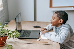 Side view of African American boy watching educational video with netbook while sitting at table with stationery
