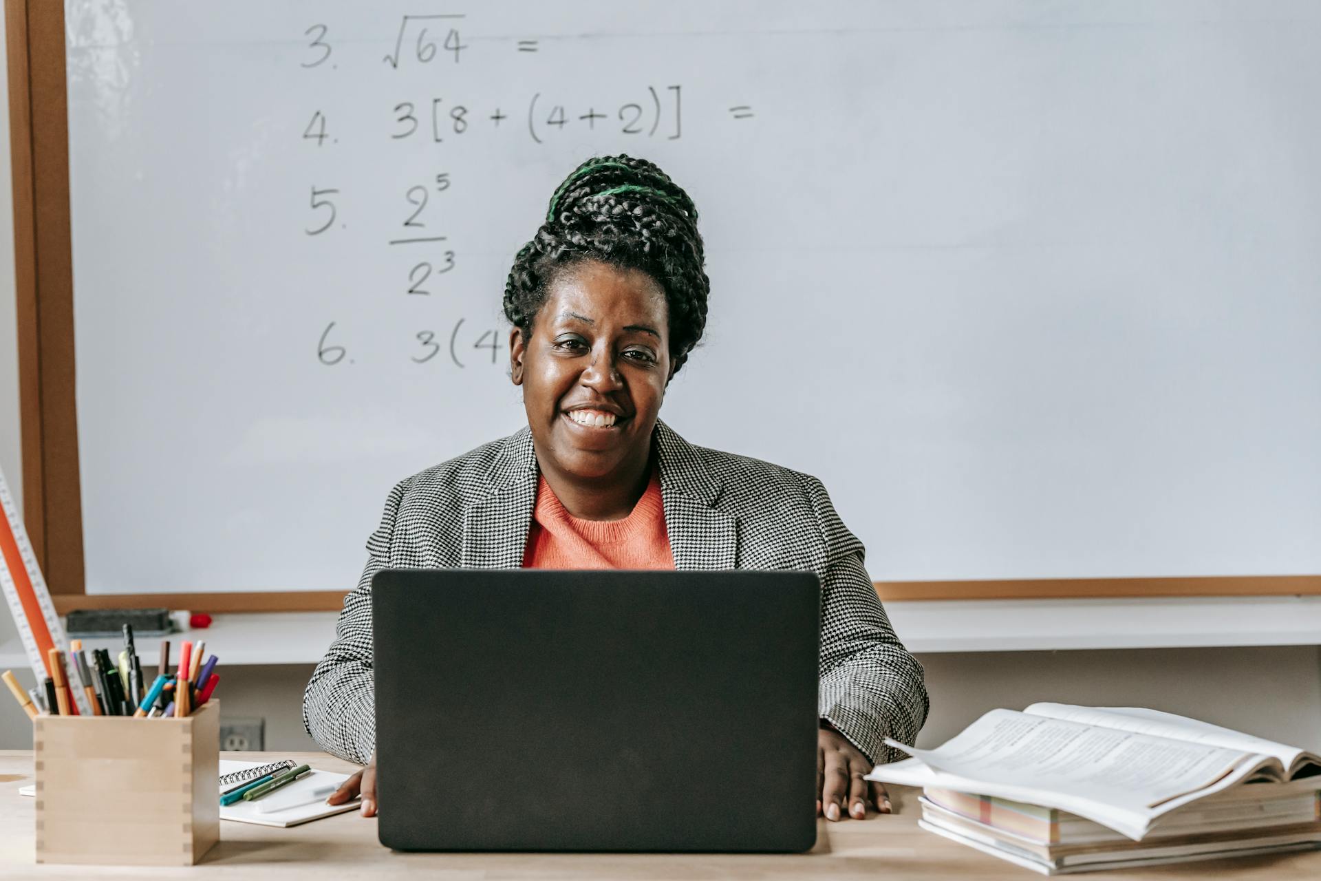 Smiling female teacher using a laptop in a classroom setting with mathematical equations on the board.