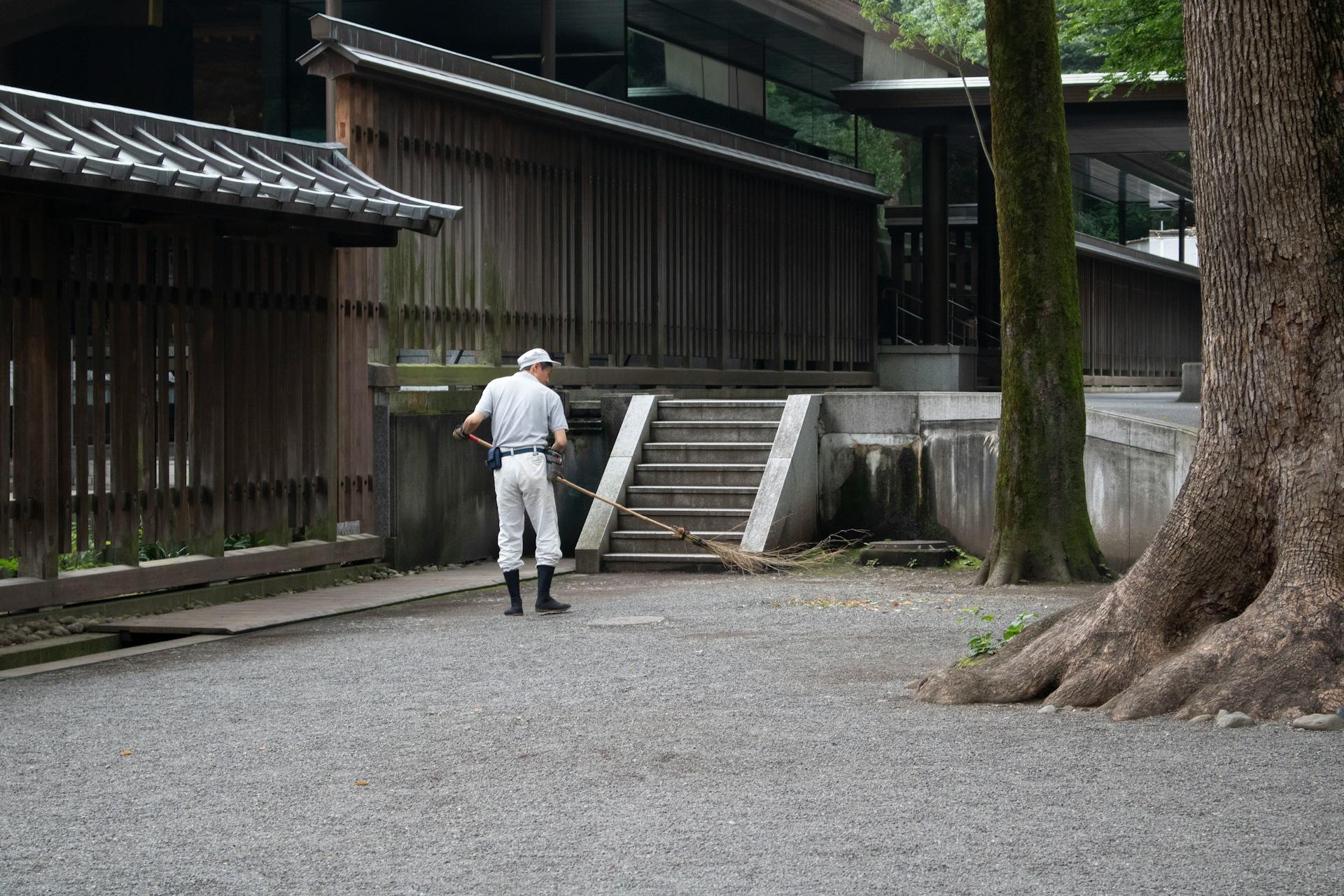 Man sweeping outdoor area near wooden building, emphasizing maintenance work.