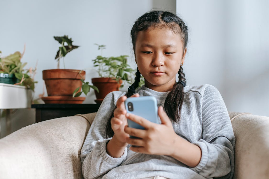 Concentrated Asian girl text messaging on smartphone while sitting on comfy armchair at home