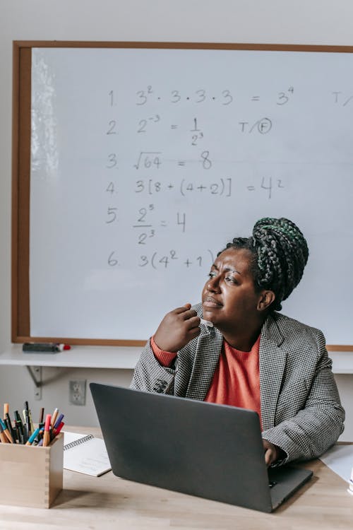 Pensive African American female teacher thinking while working on laptop in classroom and sitting at table
