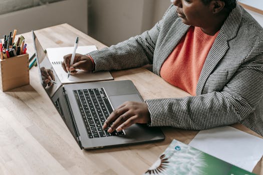 Professional woman in a formal outfit working on a laptop and writing notes at a desk.