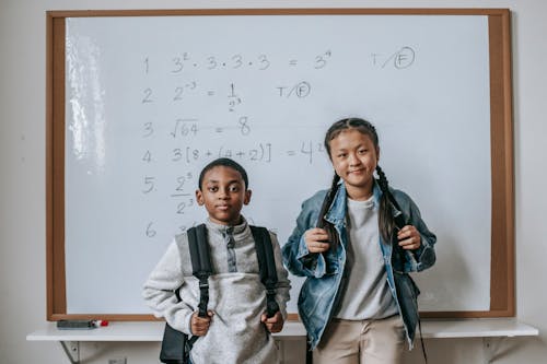 Free Focused multiracial children in casual outfits with backpacks standing in classroom against whiteboard at school and looking at camera Stock Photo