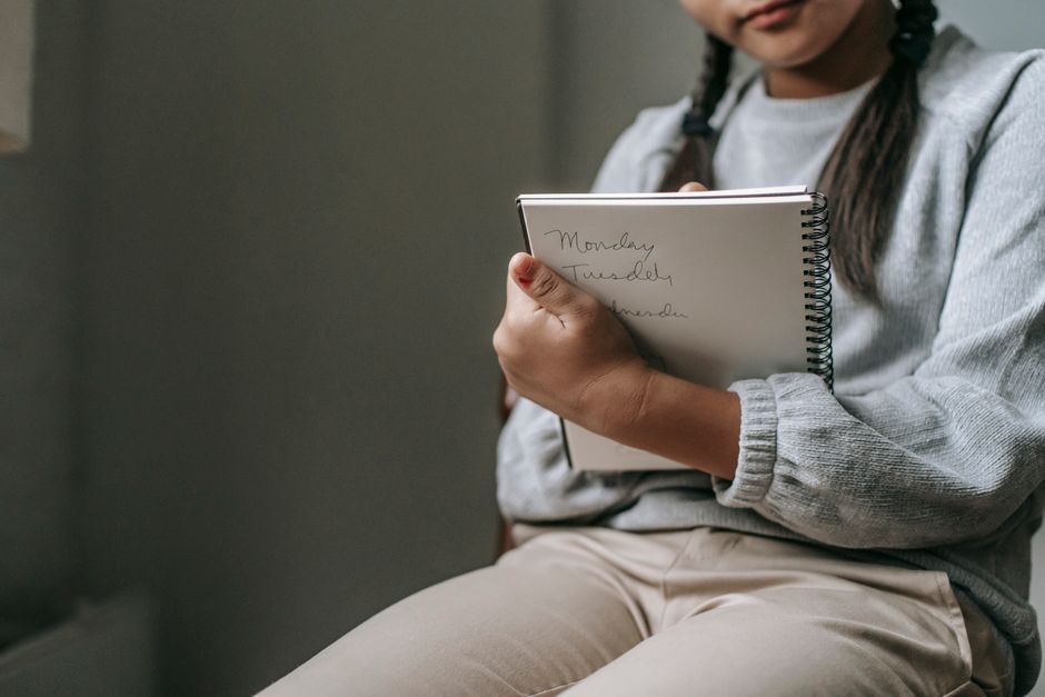 Crop unrecognizable girl in casual outfit sitting on chair and taking notes in copybook