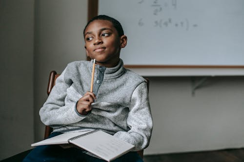 Concentrated African American boy in casual outfit sitting on chair with pencil and copybook and thinking how to do task