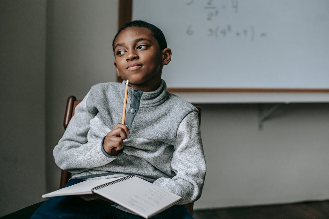 Free Concentrated African American boy in casual outfit sitting on chair with pencil and copybook and thinking how to do task Stock Photo