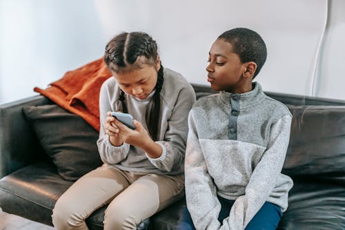 Focused Asian girl browsing internet on smartphone while curious African American boy looking at screen sitting on comfortable couch
