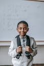 Cheerful African American boy in casual outfit with backpack standing against whiteboard in classroom and looking at camera