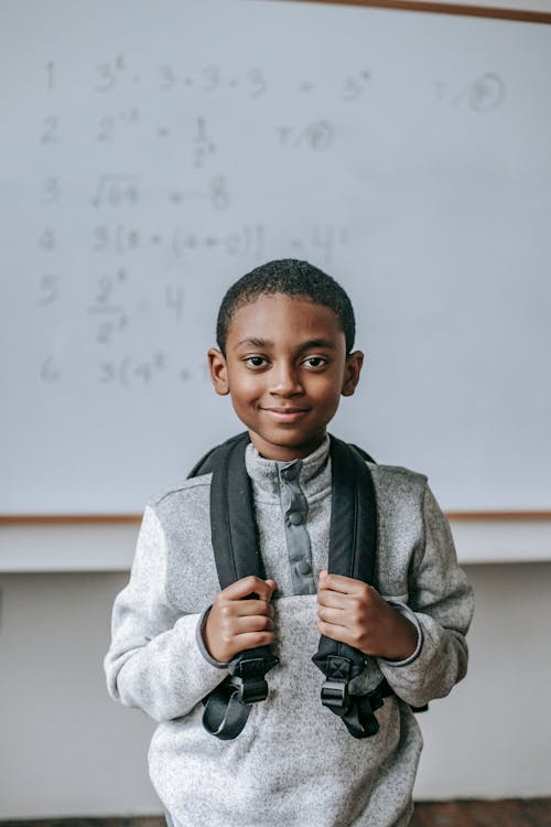 Cheerful African American boy in casual outfit with backpack standing against whiteboard in classroom and looking at camera