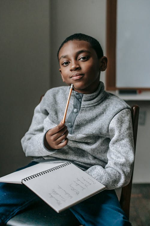 Free Focused African American boy sitting on chair and looking at camera while doing tasks in copybook Stock Photo