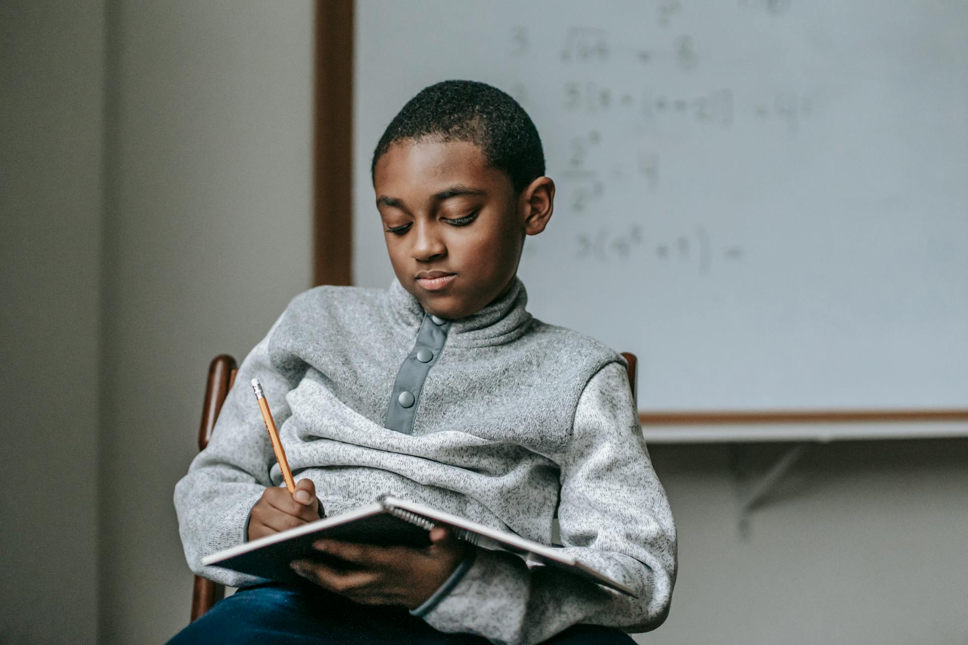 Concentrated African American boy doing tasks in copybook while sitting on wooden chair in classroom