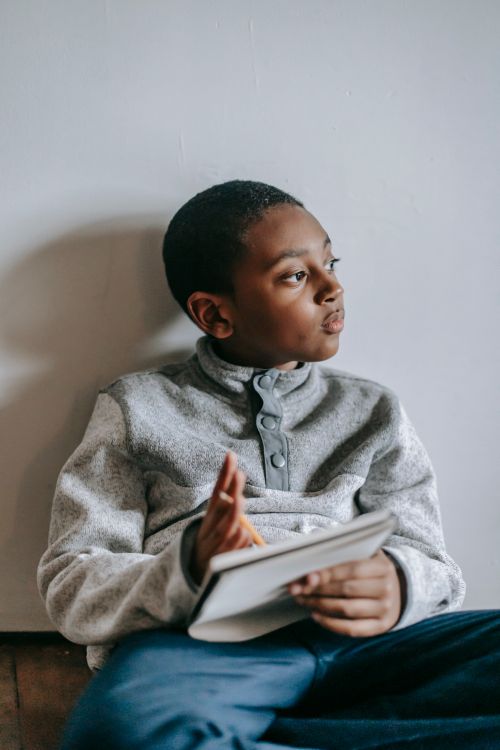Free Pensive African American schoolkid with exercise book and pencil sitting near wall while looking away Stock Photo