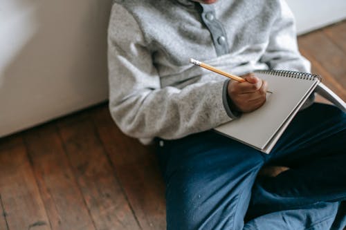 From above faceless child with pencil and notebook taking notes while sitting on floor