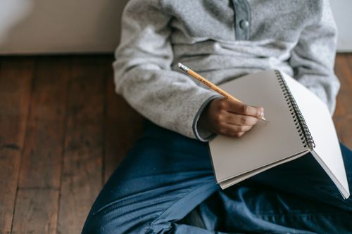 Free From above of African American schoolboy sitting on floor and writing with pencil in notebook Stock Photo