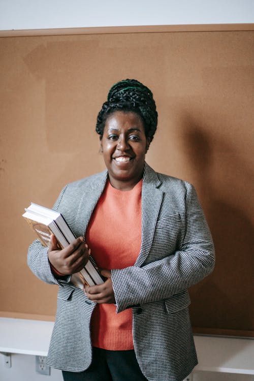 Cheerful adult black woman in jacket holding pile of books standing in classroom and looking at camera