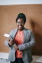 Cheerful adult black woman in jacket holding pile of books standing in classroom and looking at camera