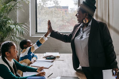 Free Side view of smiling black boy giving high five to teacher while sitting with diverse classmate girl at desk in classroom Stock Photo