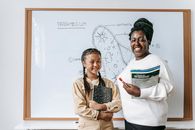 Cheerful African American female teacher near charming Asian schoolgirl with exercise book looking at camera near white board in classroom