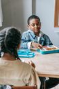 African American schoolchild with school box interacting with crop anonymous ethnic friend at table in classroom