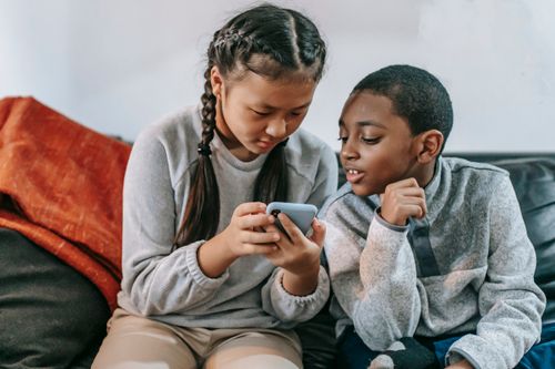Free Attentive Asian girl with black boy sharing cellphone while resting on couch in house Stock Photo