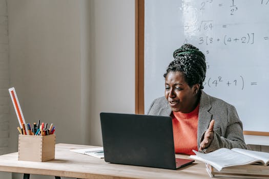 African American female teacher talking during class on netbook while sitting at desk with books and stationery in classroom