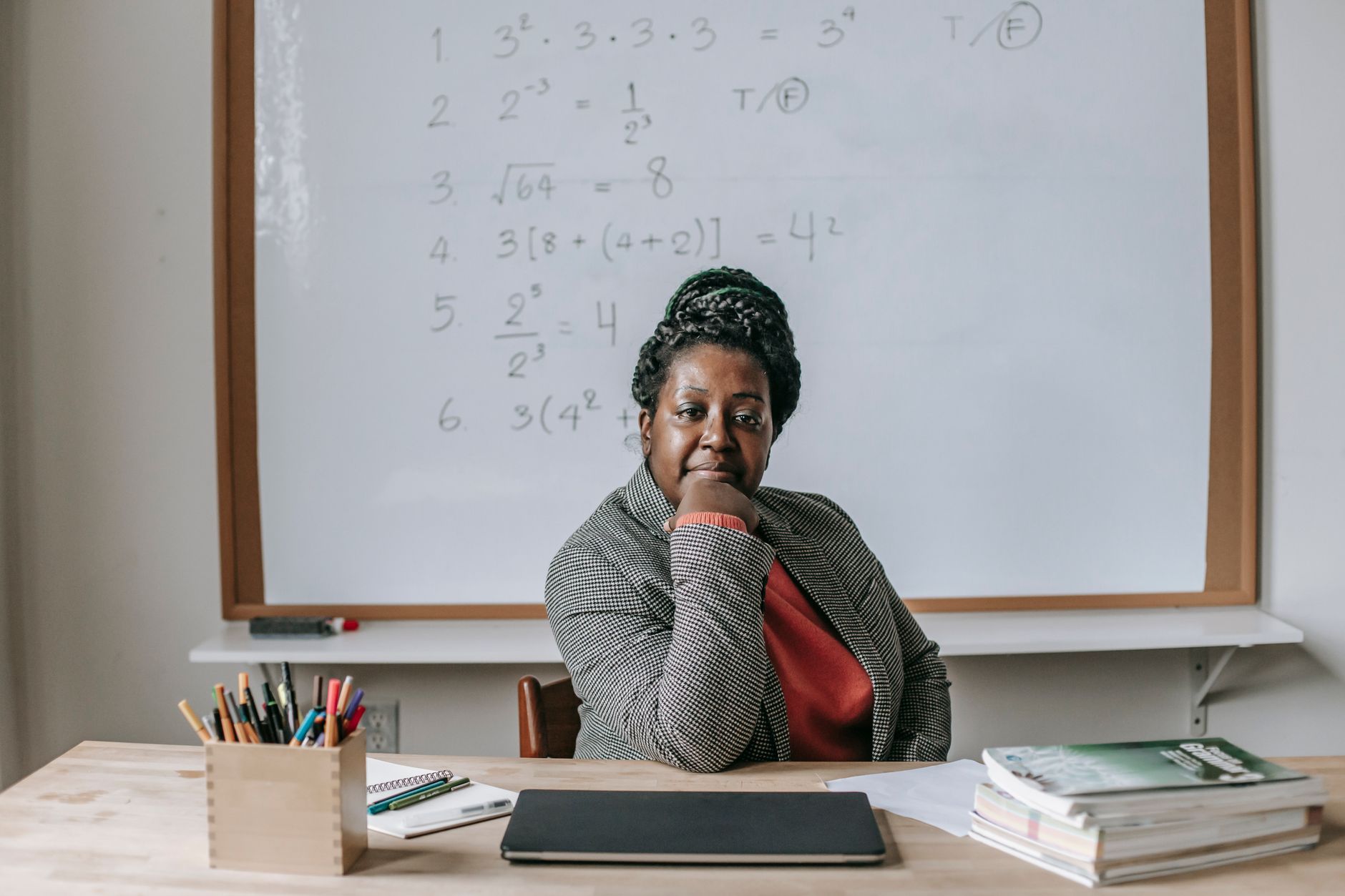 African American female teacher in formal clothes sitting at table with books and laptop and looking at camera