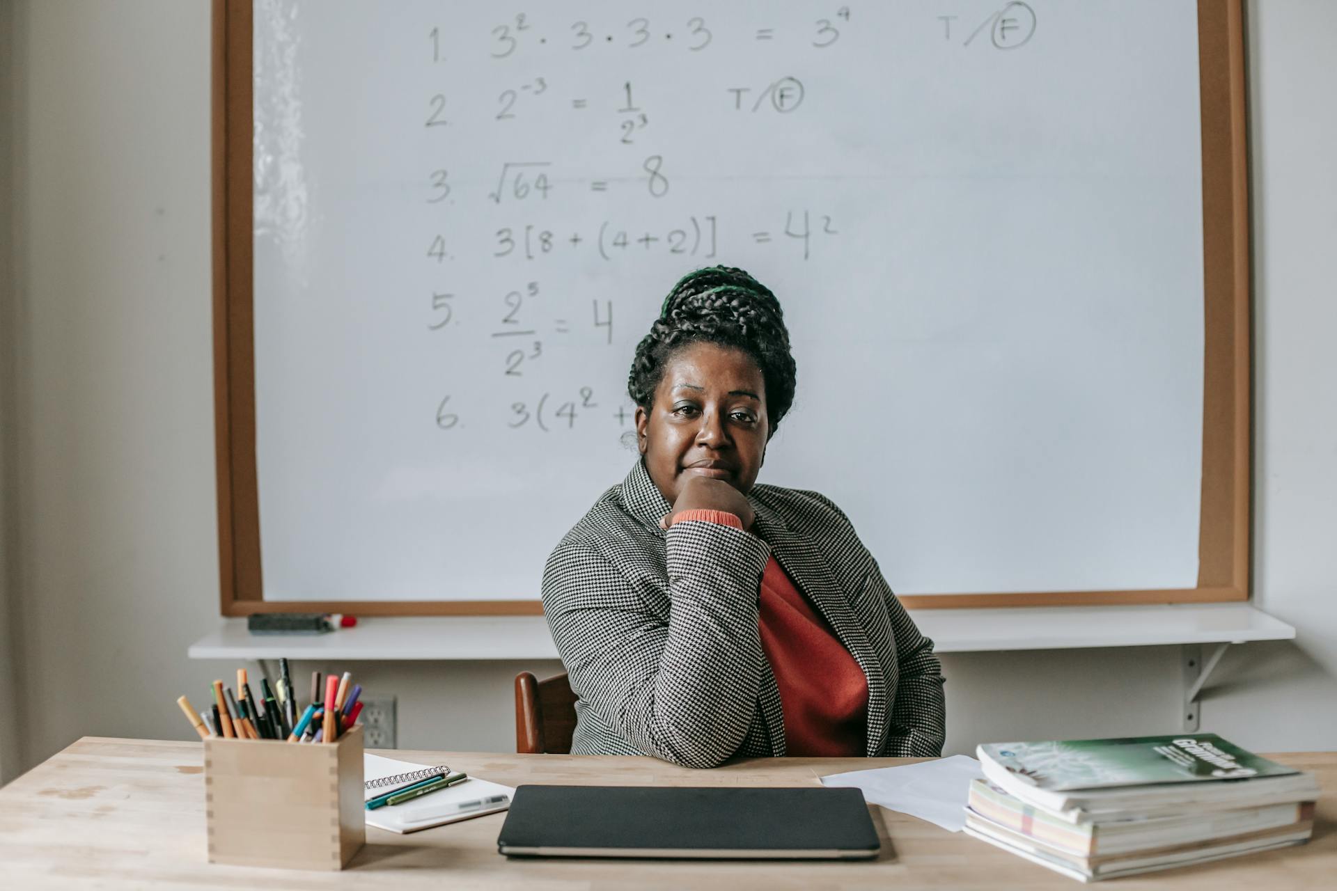 African American female teacher in formal clothes sitting at table with books and laptop and looking at camera