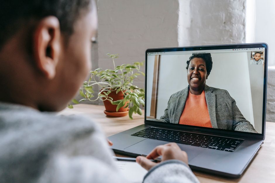 Crop anonymous African American kid sitting at table with modern laptop and listening to female teacher explaining lesson during coronavirus pandemic