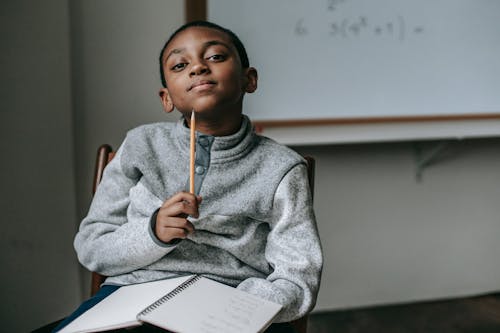 Free Cute little black boy wearing fleece sweater sitting on chair with pencil under chin and dreamy looking at camera during break after lesson in school Stock Photo