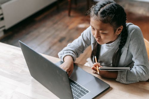 Free From above of concentrated Asian schoolgirl with pigtails sitting at wooden table with copybook and pencil and browsing laptop for school task Stock Photo