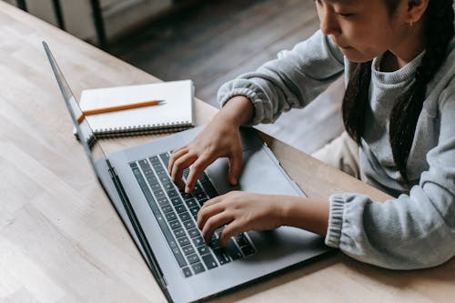 Crop adorable schoolgirl typing on wireless laptop at wooden desk