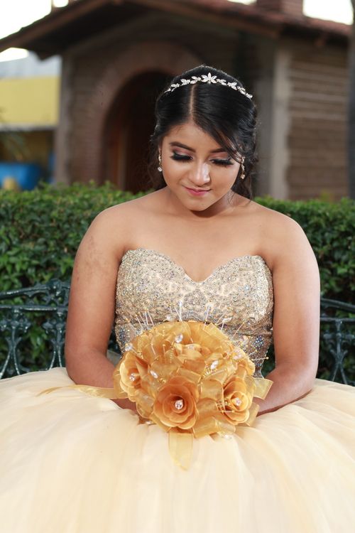 Free A graceful woman in a glamorous gold gown with a tiara, holding a vibrant bouquet outdoors. Stock Photo