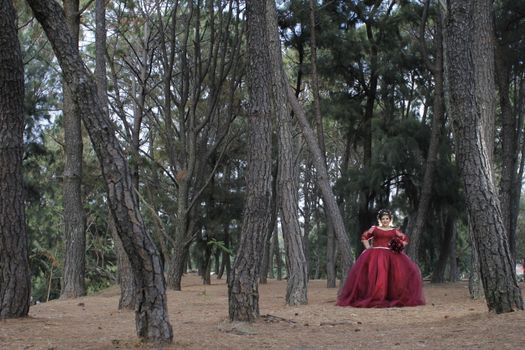 Elegant woman in a flowing red dress holding a bouquet, standing in a serene forest setting.