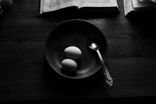 From above black and white of chicken eggs with spoon in round shaped bowl near open books at home