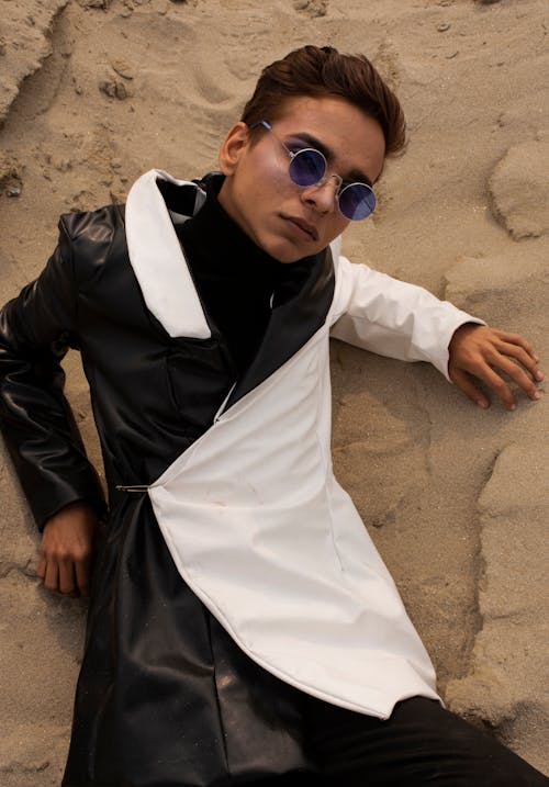 Man in Black and White Coat Wearing Sunglasses Lying Down on Sand