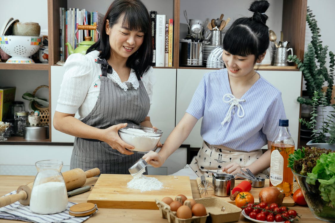 Smiling Asian mother wearing apron standing near wooden counter and watching daughter mixing ingredients for dough in light modern kitchen