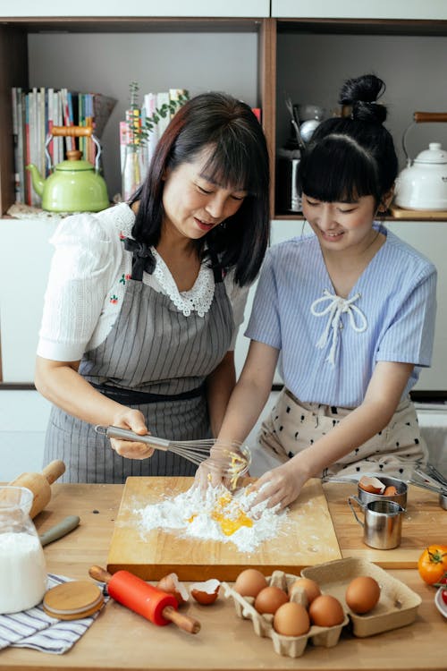 Delightful Asian woman and daughter mixing ingredients for dough together in kitchen