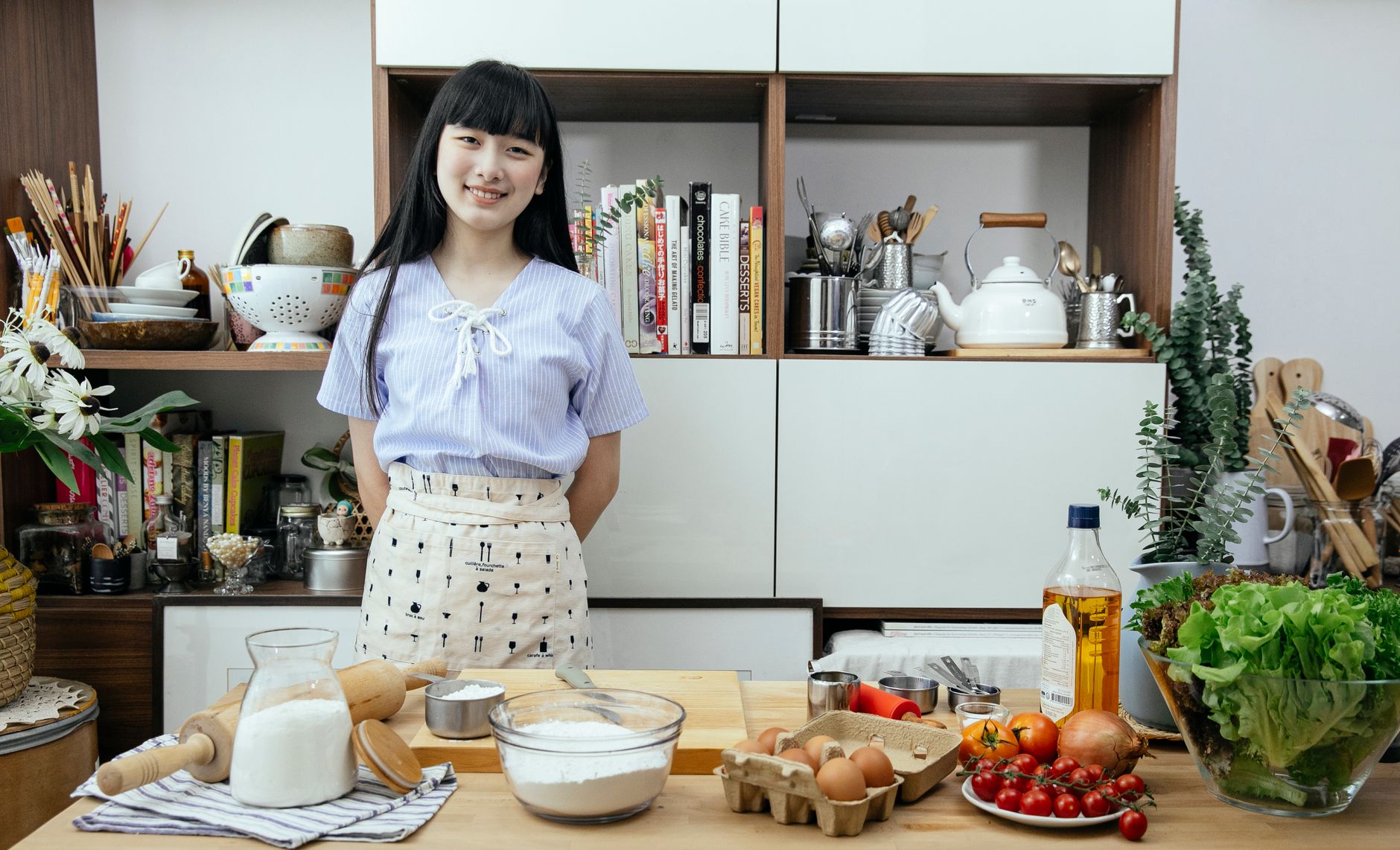 Smiling young Asian lady wearing apron standing near wooden counter with various vegetables and ingredients for dough in light modern kitchen