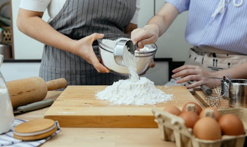 Crop anonymous woman in apron and young lady standing near counter and pouring flour on wooden cutting board in modern kitchen