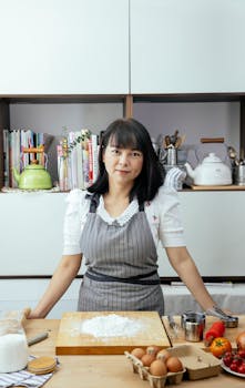 Positive adult Asian woman wearing apron leaning on wooden table with eggs and tomatoes for cooking in contemporary light kitchen at home