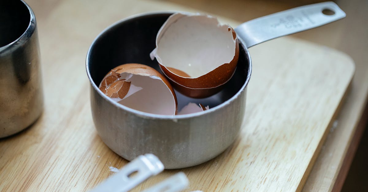 From above of stainless steel scoop with crashed eggshells placed on wooden table in kitchen in daylight